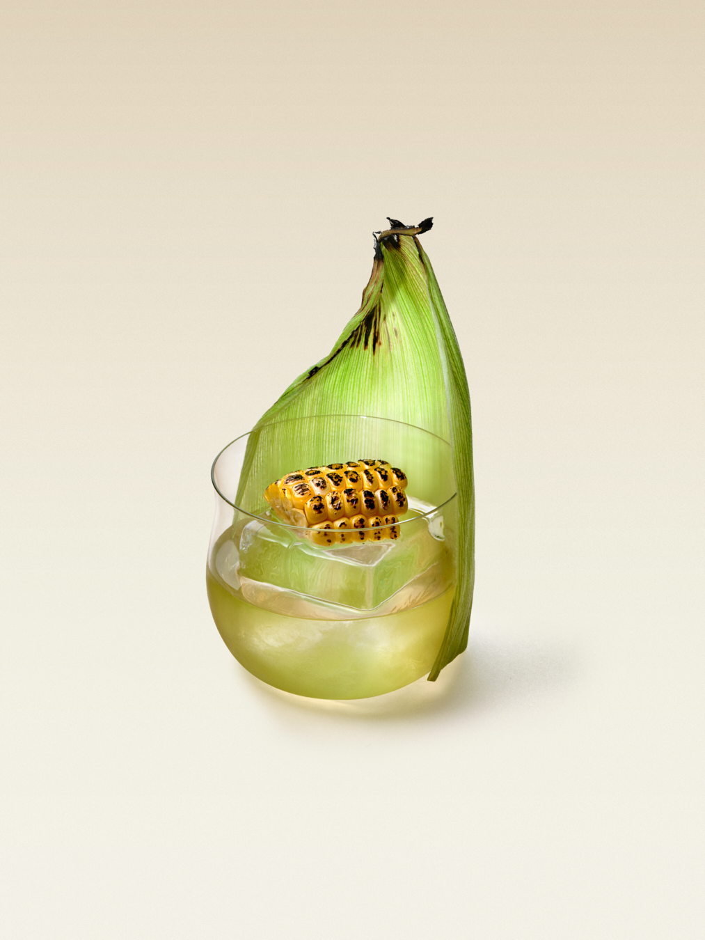 Short cocktail glass with one large ice cube and husk of corn on the side of the glass, displaying an Elote Nixta Corn Liqueur cocktail. 