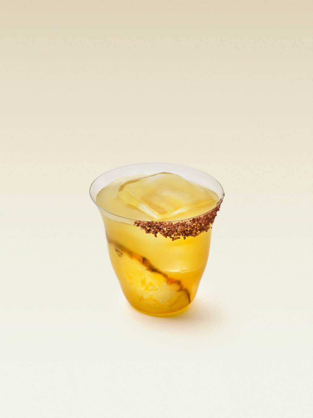 A short glass with one large square ice cube, the glass rimmed with orange chile salt and one slice of pineapple inside the glass, displaying a spicy pineapple margarita.
